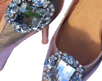 May include: A pair of gold satin high heels with a pointed toe and a large, square-shaped rhinestone embellishment on the front of each shoe.