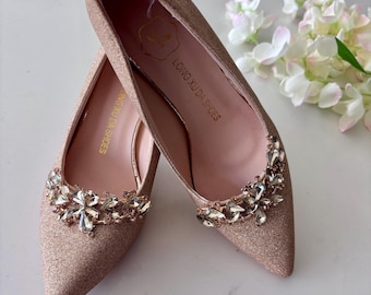 May include: A pair of pointed-toe flats in a rose gold glitter finish. Each shoe features a decorative cluster of clear, faceted crystals on the toe. The interior insole is light pink and printed with the text LONG XU DA SHOES in gold lettering.