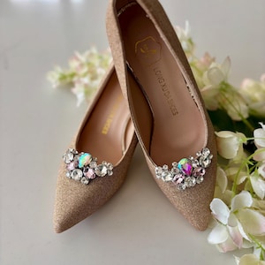 Puede incluir: Un par de zapatos de tacón dorados con punta y adornos de joyas. Los zapatos de tacón bajo presentan un grupo de cristales transparentes e iridiscentes. La marca "LONG XU DA SHOES" está impresa en el interior.