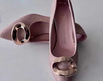 May include: A pair of rose gold glitter pointed-toe pumps with a large gold-tone buckle detail. The shoes have a stiletto heel and a shiny finish, perfect for a special occasion. The interior of the shoe is a light pink color.