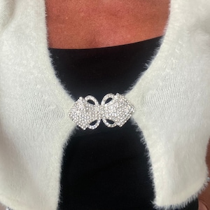 Pode incluir: Um casaco branco felpudo com um grande broche de laço de strass prateados.
