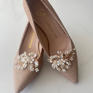 Puede incluir: Un par de zapatos de tac&oacute;n alto de brillo dorado con un adorno floral en la punta. El adorno presenta flores blancas, hojas doradas y cuentas de perlas. Los zapatos est&aacute;n etiquetados como "LONG XU DA SHOES".