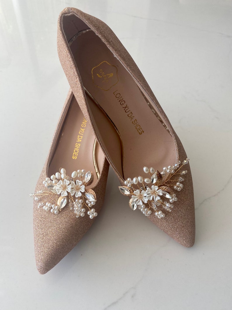 Puede incluir: Un par de zapatos de tac&oacute;n alto de brillo dorado con un adorno floral en la punta. El adorno presenta flores blancas, hojas doradas y cuentas de perlas. Los zapatos est&aacute;n etiquetados como "LONG XU DA SHOES".