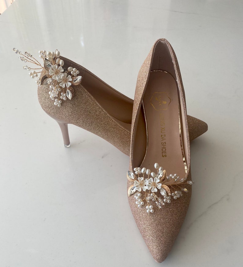Puede incluir: Un par de tacones altos de brillo dorado con punta puntiaguda y un delicado adorno floral en cada zapato. Los adornos presentan flores blancas, perlas y detalles dorados. Los zapatos est&aacute;n etiquetados como "Xing Xuda Shoes".