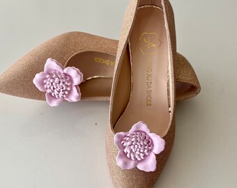May include: A pair of gold glitter pointed-toe heels with a pink flower embellishment on each shoe. The shoes have the text "Xing Xu Da Shoes" on the inside of one shoe.