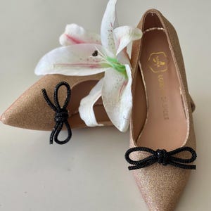 Op de afbeelding: Een paar gouden glitter spitsschoenen met zwarte strikjes. De schoenen staan op een witte ondergrond met een witte lelie.