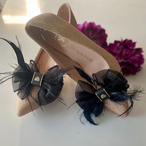 Può includere: Un paio di scarpe dorate con glitter, fiocchi neri e piume. Le scarpe hanno il testo "LONG XU DA SHOES" all'interno.