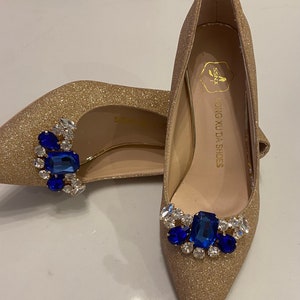 以下が含まれることがあります： 青いラインストーンとクリアなラインストーンで飾られた、ゴールドのグリッターハイヒール。靴はポインテッドトゥとスティレットヒール。靴の中敷きには「Hong Xuda Shoes」と書いてあります。