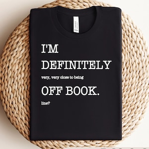 May include: Black t-shirt with white text that reads "I'M DEFINITELY very, very close to being OFF BOOK. line?" The shirt is displayed on a woven, circular surface.