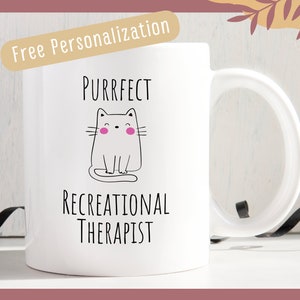 May include: White ceramic mug with a black and white illustration of a cat with pink cheeks. The text "Purrfect Recreational Therapist" is printed on the mug.