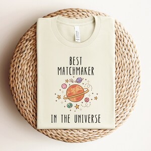 May include: A light beige t-shirt with the text "Best Matchmaker in the Universe" and a cartoon illustration of a planet with stars and other planets around it.