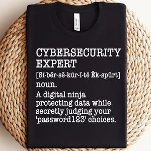 May include: Black t-shirt with white text that reads "CYBERSECURITY EXPERT [Si-běr-sě-kür-i-tē Ěk-spûrt] noun. A digital ninja protecting data while secretly judging your 'password123' choices."