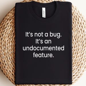 May include: Black t-shirt with white text that reads "It's not a bug. It's an undocumented feature."