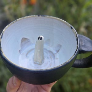May include: A ceramic mug with a matte black exterior and a glossy white interior. A small, sculpted white ghost figure with two black dots for eyes is attached to the center of the interior base, rising upward from the bottom of the mug.