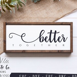 Op de afbeelding: Een rechthoekig houten bord met de woorden "better together" in zwarte letters op een witte achtergrond. Het bord is omlijst met donkerbruin hout. Het bord ligt op een jute oppervlak met decoratieve elementen.