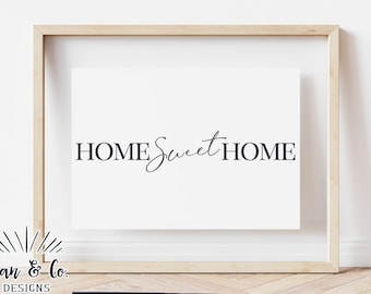 Op de afbeelding: Een ingelijst afdruk met de tekst "HOME Sweet HOME" in een eenvoudig, modern lettertype. De afdruk wordt weergegeven op een witte muur met een houten vloer.