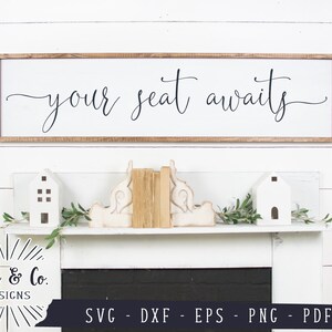 May include: A white wooden sign with the text "your seat awaits" in black cursive script. The sign is hanging on a white wall with white wood paneling. The sign is above a white mantel with a black fireplace.