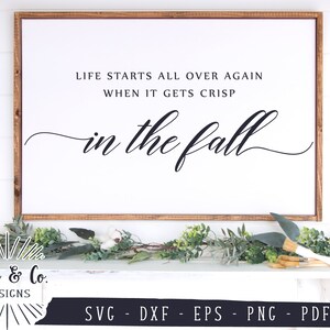 May include: A white sign with a brown wood frame, featuring the text "Life starts all over again when it gets crisp in the fall". The sign is sitting on a white surface with green foliage.