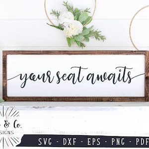May include: A wooden sign with a white background and brown frame. The sign says "your seat awaits" in black cursive lettering.