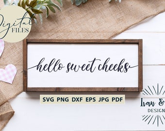 May include: A white wooden sign with the text "hello sweet cheeks" in black cursive font. The sign is on a white wooden background with a burlap texture in the top left corner. The sign is surrounded by greenery and pink gingham hearts.
