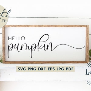 May include: A white sign with a wooden frame, featuring the text "HELLO pumpkin" in black cursive script. The sign is sitting on a white surface with a green plant in the foreground.