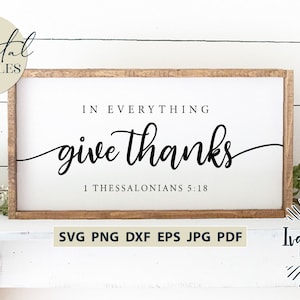 Peut inclure: Une pancarte en bois avec le texte "IN EVERYTHING give thanks 1 THESSALONIANS 5:18" en écriture cursive noire. La pancarte est posée sur une surface blanche avec des plantes vertes de chaque côté.