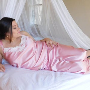 May include: A light pink satin pajama set, including a camisole with lace trim and matching pants. The model is wearing navy blue heels. The set is displayed on a white bed with a sheer canopy in the background.