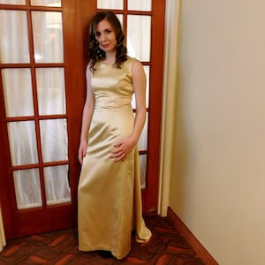 May include: A floor-length, sleeveless, golden-yellow satin gown. The dress features a fitted bodice and a flowing skirt with a slight train. The woman is standing in front of a dark wood door with glass panels.