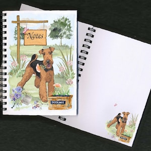 Può includere: Un quaderno a spirale con un'illustrazione ad acquerello di un cane Airedale Terrier marrone e nero in un ambiente da giardino. Il cane tiene un fiore in bocca. La copertina presenta anche un cartello con la scritta "Notes".
