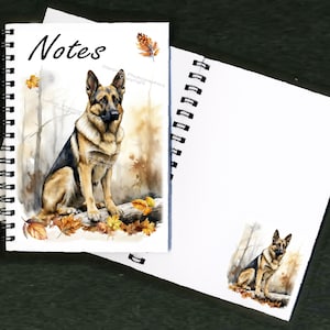 Puede incluir: Un cuaderno de espiral con una pintura de acuarela de un pastor alemán sentado en un tronco en un entorno de bosque. El perro es marrón y negro con un pecho blanco. El fondo es una pintura de acuarela de un bosque con hojas de otoño. La portada del cuaderno dice "Notes".