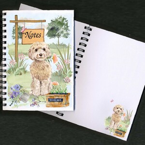 May include: A spiral bound notebook with a watercolor illustration of a light brown dog sitting in a garden setting. The dog is holding a flower in its mouth. The front cover of the notebook has the word "Notes" written on a sign hanging from a wooden post. The back cover of the notebook is blank.