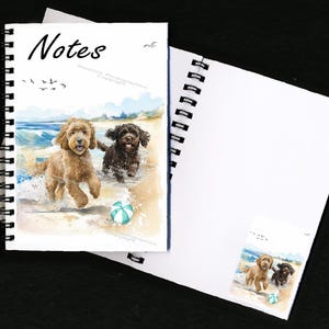 Puede incluir: Un cuaderno de espiral con una ilustración de acuarela de dos perros jugando en la playa. Los perros son un doodle marrón y blanco y un doodle negro y marrón. La portada del cuaderno dice "Notes" en la parte superior y tiene un aviso de copyright en la parte inferior.