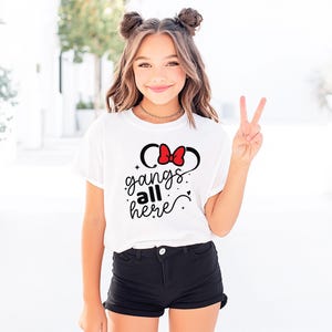 K&ouml;nnte beinhalten: Wei&szlig;es T-Shirt mit roter Schleife und schwarzem Mauseohren-Design sowie dem Text "gangs all here" in Schreibschrift. Das Shirt wird mit schwarzen Shorts kombiniert. Die Person, die das Shirt tr&auml;gt, zeigt das Victory-Zeichen.