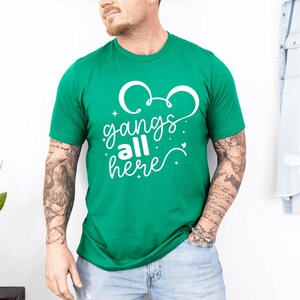 K&ouml;nnte beinhalten: Ein gr&uuml;nes T-Shirt mit wei&szlig;em Text, der "gangs all here" lautet. Das Design enth&auml;lt eine Mickey Mouse-Ohrenform &uuml;ber dem Text. Das Shirt hat eine einfarbige gr&uuml;ne Farbe.