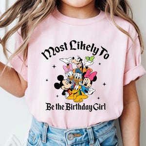 May include: A light pink t-shirt featuring a graphic of Disney characters including Mickey Mouse, Minnie Mouse, Donald Duck, Daisy Duck, Goofy, and Pluto. The text "Most Likely To Be the Birthday Girl" is printed above and below the characters.