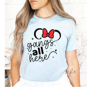 K&ouml;nnte beinhalten: Hellblaues T-Shirt mit einem Minnie-Mouse-inspirierten Design. Das Design zeigt schwarze Micky-Maus-Ohren mit einer roten Schleife und den Schriftzug "gangs all here" in einer verspielten Schriftart. Ein l&auml;ssiges Kleidungsst&uuml;ck.