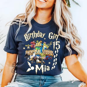 May include: Navy blue t-shirt with a graphic featuring the text "Birthday Girl 15" and "Universal Studios Mia." The design includes various cartoon characters and a footprint trail.