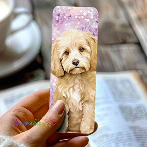 Può includere: Un segnalibro con un carino cane marrone chiaro dal pelo soffice. Il cane è seduto su uno sfondo rosa e bianco con una texture scintillante.