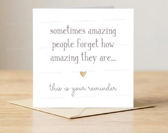 May include: A white greeting card with the text "sometimes amazing people forget how amazing they are..." and "this is your reminder" in a handwritten style. A small gold heart is below the text. The card rests on a brown envelope.