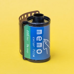 May include: A roll of Memo colour film with a blue label and white text. The label includes the text "memo colour film" and "200 iso 36 exp". The film canister is black with a yellow background.
