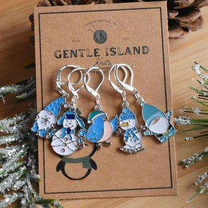 May include: Set of five blue and white enamel stitch markers featuring a snowman, a gnome, a bird, and two more snowmen. The markers are attached to silver jump rings. The card reads "Gentle Island Knits".