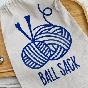 May include: A beige canvas ball sack with a blue yarn ball design and the words "BALL SACK" printed in blue. Knitting accessories, including stitch markers and a knitting needle, are on a wooden tray.