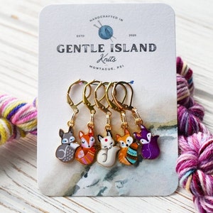 May include: Set of five fox-themed stitch markers with gold-toned hoops. The foxes are painted in various colours and patterns, including purple, orange, white, and grey. The markers are displayed on a card with the text "Gentle Island Knits, Montague, PEI".