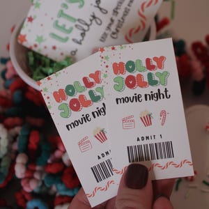 May include: Two "Holly Jolly Movie Night" tickets with festive graphics and a barcode. The tickets are held in front of a white container with a "Let's Watch a holly jolly movie!" card.