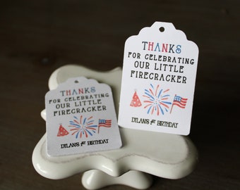 Puede incluir: Dos etiquetas de regalo blancas con diseños rojo, blanco y azul. El texto en las etiquetas dice "Gracias por celebrar nuestro pequeño petardo" y "Dilans 1er cumpleaños".