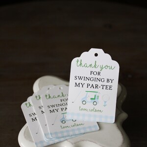 May include: A stack of white thank you tags with blue and white checkered pattern on the bottom. The tags have green text that says "thank you for swinging by my par-tee" with a cartoon golf cart and golf ball. The text "love, colson" is printed on the bottom of the tag.