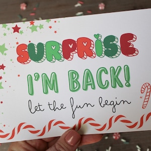 May include: A white card with the words "SURPRISE I'M BACK!" in green and red bubble letters. The phrase "let the fun begin" is written in cursive. The card is decorated with stars and candy cane accents.