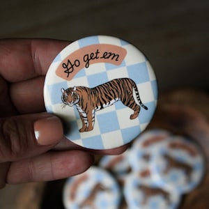 May include: A round button with a tiger illustration. The tiger is brown and black, set against a blue and white checkered background. The text "Go get em" is in a cursive font. Additional buttons are visible.