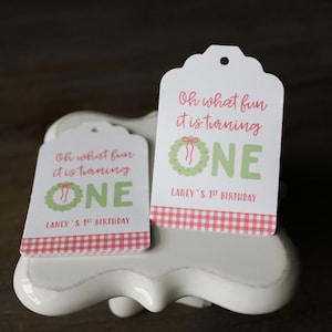 Puede incluir: Dos etiquetas de regalo blancas con bordes festoneados. Las etiquetas tienen el texto "Oh what fun it is turning ONE" en rojo y verde, con un diseño de corona. La parte inferior de las etiquetas tiene un patrón de cuadros rojos y blancos. También está presente el texto "Laney's 1st Birthday".