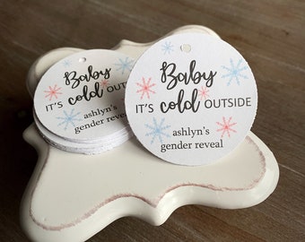 May include: Two white round paper tags with blue and pink snowflakes and the text "Baby cold outside" and "It's ashln's gender reveal".